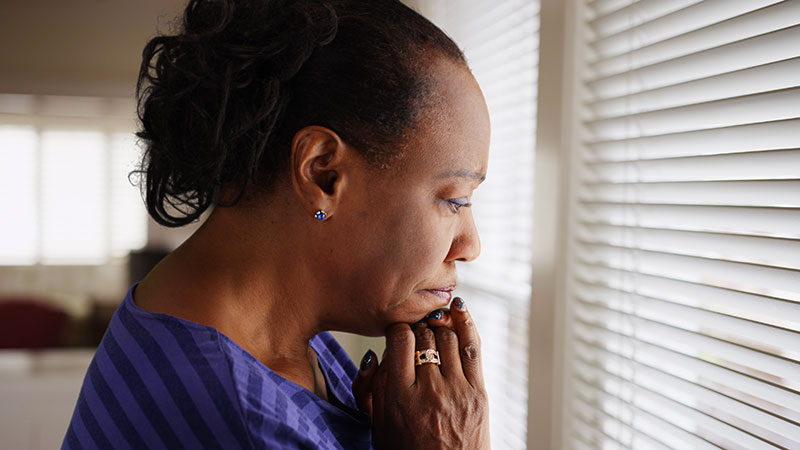 elderly black woman sadly gazing out a window with blinds