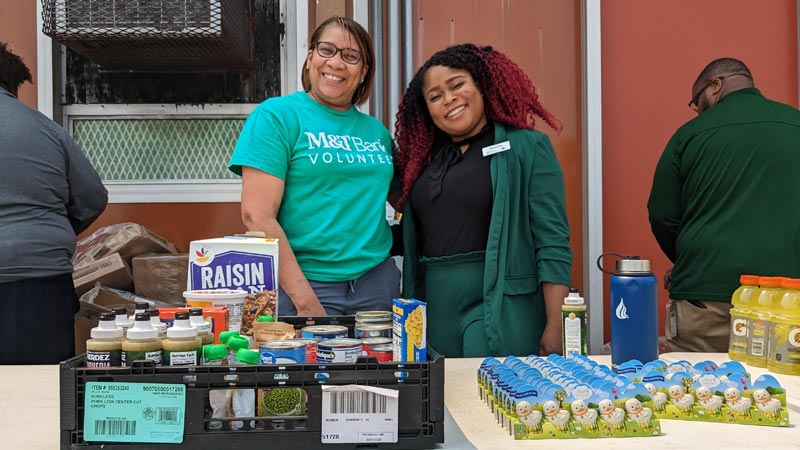 two smiling women standing outside at food distribution table