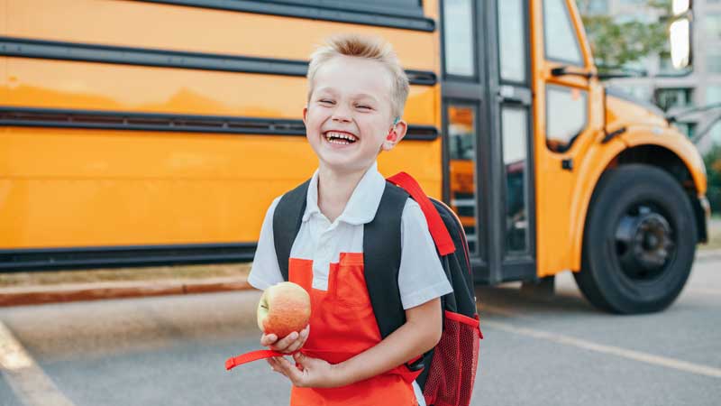 elementary school boy laughing in front of a school bus holding an apple
