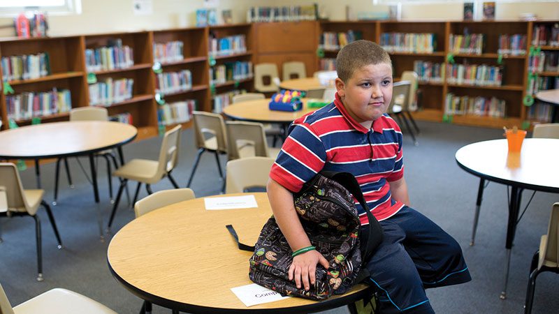 young caucasian boy sitting in school library with his backpack