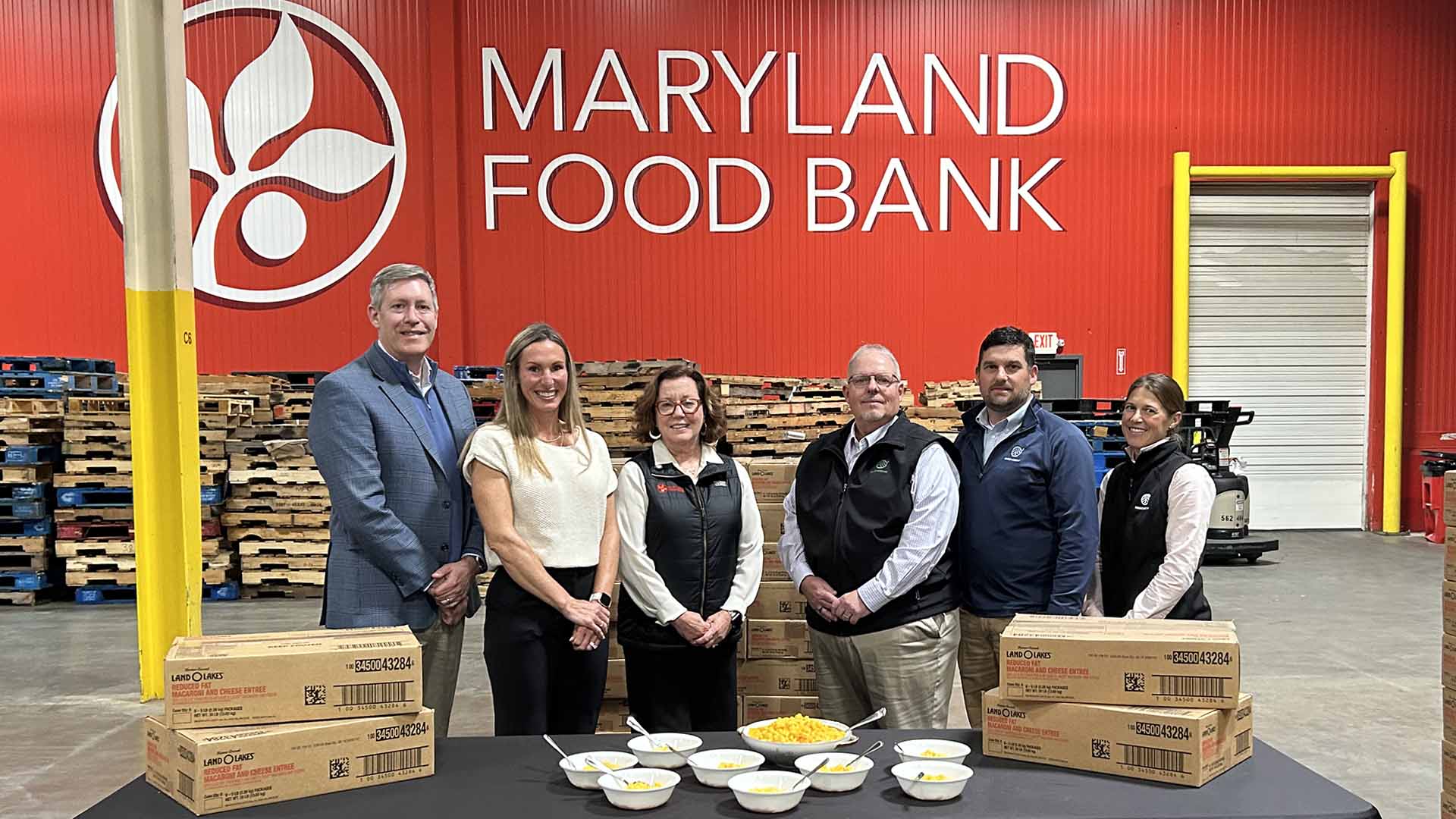 group of representatives from Maryland Department of Agriculture, Maryland Food Bank, USDA, Land O' Lakes standing behind a table with boxes and bowls of macaroni and cheese in front of a red wall with the Maryland Food Bank logo in a warehouse