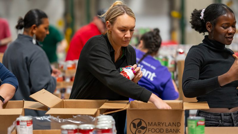 caucasian female volunteer standing beside a black female volunteer packing holiday meal kit boxes