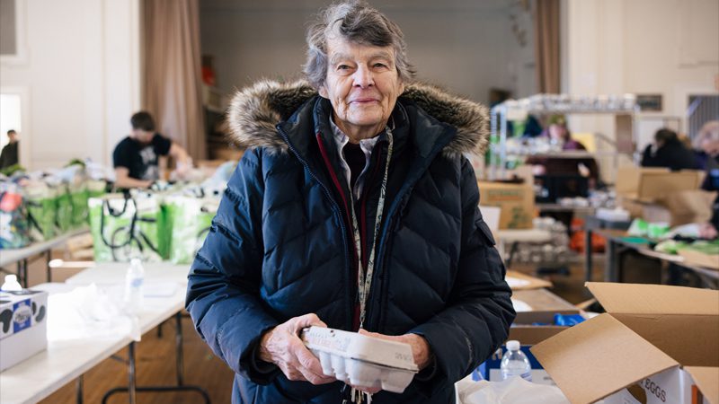 elderly woman stands at food pantry in winter coat holding a carton of eggs