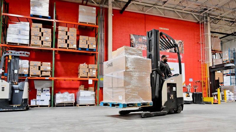 male Maryland Food Bank employee operating forklift carrying wrapped pallet of boxed food labeled as snacks