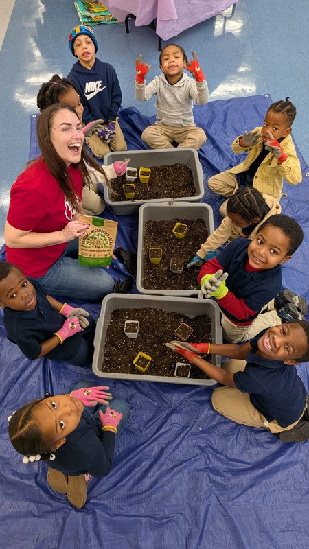 Ashlie Church kneels on a blue tarp with elementary students to teach them how to garden in plastic bins