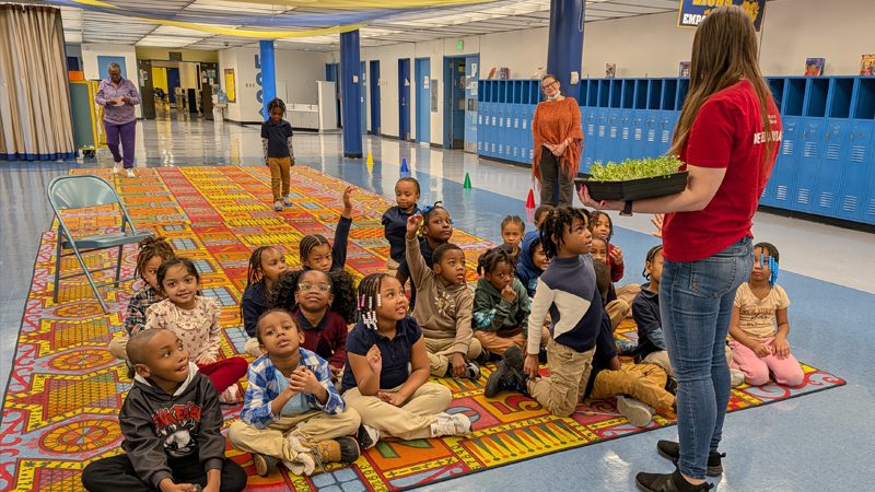 Ashlie Church stands in elementary school teaching a class who is sitting on a multi-colored rug
