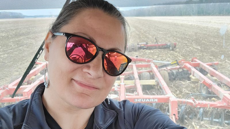 Nicole Wickham smiles in her sunglasses while driving a tractor picking crops