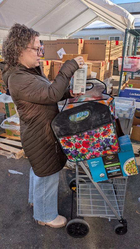 middle-aged caucasian female in brown winter coat adding groceries to her cart at outside an Open Hands food distribution