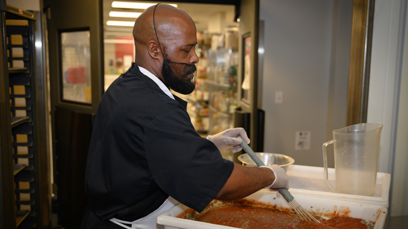 Eric Latta in a black shirt and beard mask stirs a large container of spaghetti sauce using a large wisk