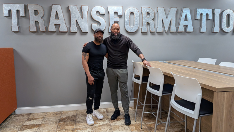 two men standing in front of a wall with large letters spelling TRANSFORMATION